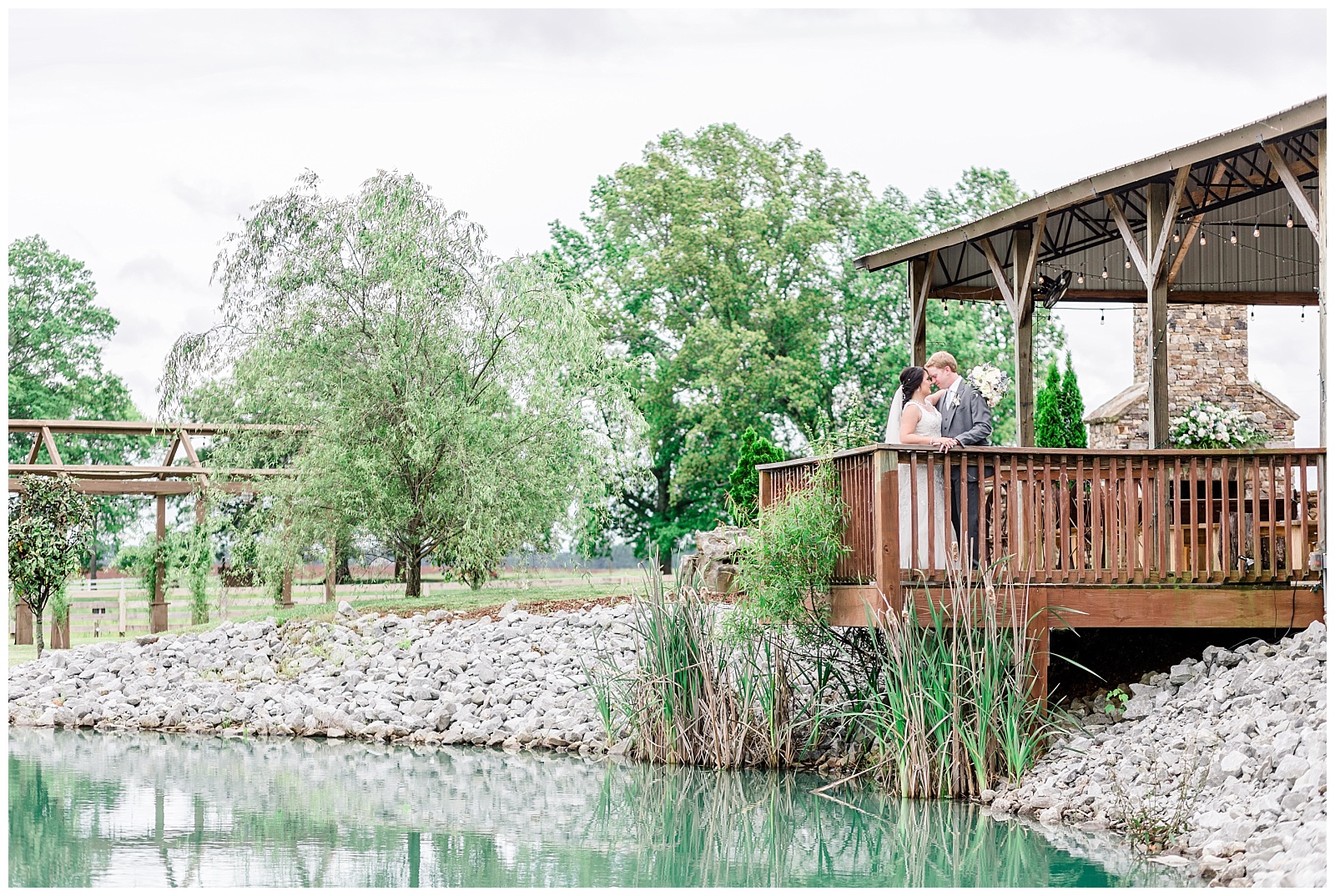 Rustic Farm Alabama Wedding in front of southern pond with mermaid style wedding dress, flowy beaded veil, grey suit, and white, blue, yellow, and pink florals