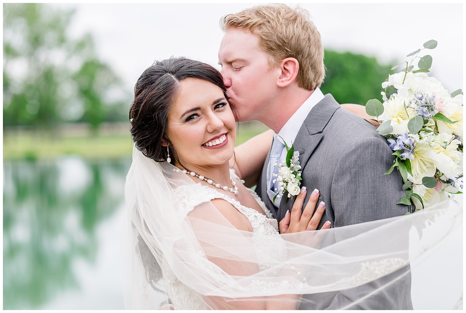 Rustic Farm Alabama Wedding in front of southern pond with mermaid style wedding dress, flowy beaded veil, grey suit, and white, blue, yellow, and pink florals